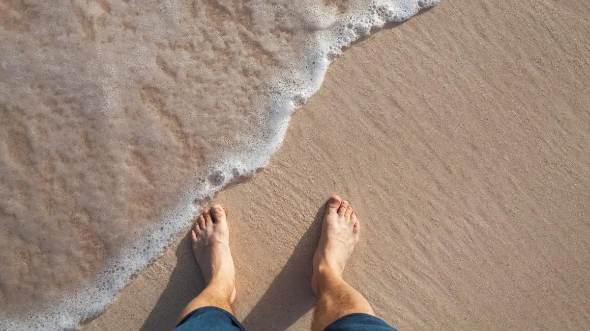 Man standing barefoot on wet sand with waves washing in, illustrating grounding, sensory feedback and foot awareness